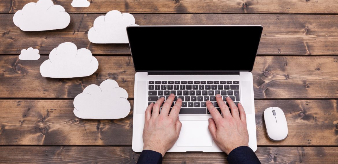 Cloud computing technology concept with white fluffy clouds next to the laptop. Mans hands typing the the keyboard uploading data, on a wooden table.